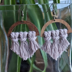 May include: A pair of wooden hoop earrings with a light purple tassel fringe. The hoops are made of dark wood and have a semi-circular shape. The tassels are made of a soft, light purple yarn and are tied in a knot at the top.