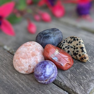 May include: Five tumbled stones in various colors and patterns, including a dark gray stone, a light orange stone, a red stone, a purple stone, and a black and white speckled stone.