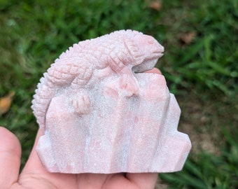 May include: A pink stone carving of a lizard perched on a rocky outcrop. The carving is detailed and intricate, with a textured surface.