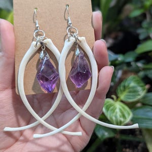 Peut inclure: Boucles d'oreilles faites à la main avec des accents d'os et des pendentifs en cristal violet facetté. Les boucles d'oreilles ont des crochets argentés et sont présentées sur une carte marron. Le design s'inspire de la nature et de l'esthétique gothique.