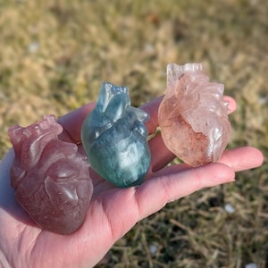 Puede incluir: Tres esculturas de corazón de resina en diferentes colores: rosa, azul y transparente con vetas rojas. Los corazones se muestran en una mano.