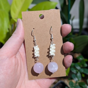 May include: A pair of dangle earrings featuring a bone-like design and light purple spherical beads. The earrings are attached to silver-colored hooks and are displayed on a brown card.