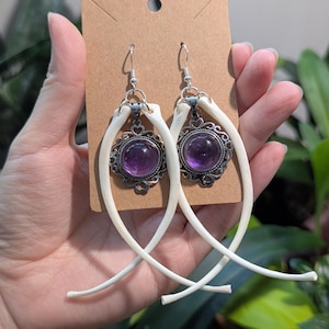 Peut inclure: Une paire de boucles d'oreilles uniques avec une pierre précieuse violette sertie dans un cadre argenté orné. Les boucles d'oreilles sont suspendues à un crochet argenté et sont accentuées d'éléments incurvés en forme d'os. Présentées sur une carte brune.