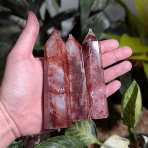 Könnte beinhalten: Drei rote und klare Quarzkristallspitzen, auch bekannt als Zauberstäbe, werden in einer Hand gehalten. Die Kristalle sind durchscheinend und haben eine rotbraune Farbe.
