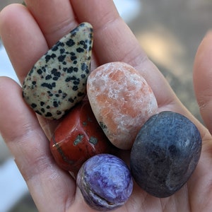 May include: A collection of five polished stones in various colors and patterns, including a black and white speckled stone, a red stone, a light orange stone, a dark blue stone, and a purple stone.