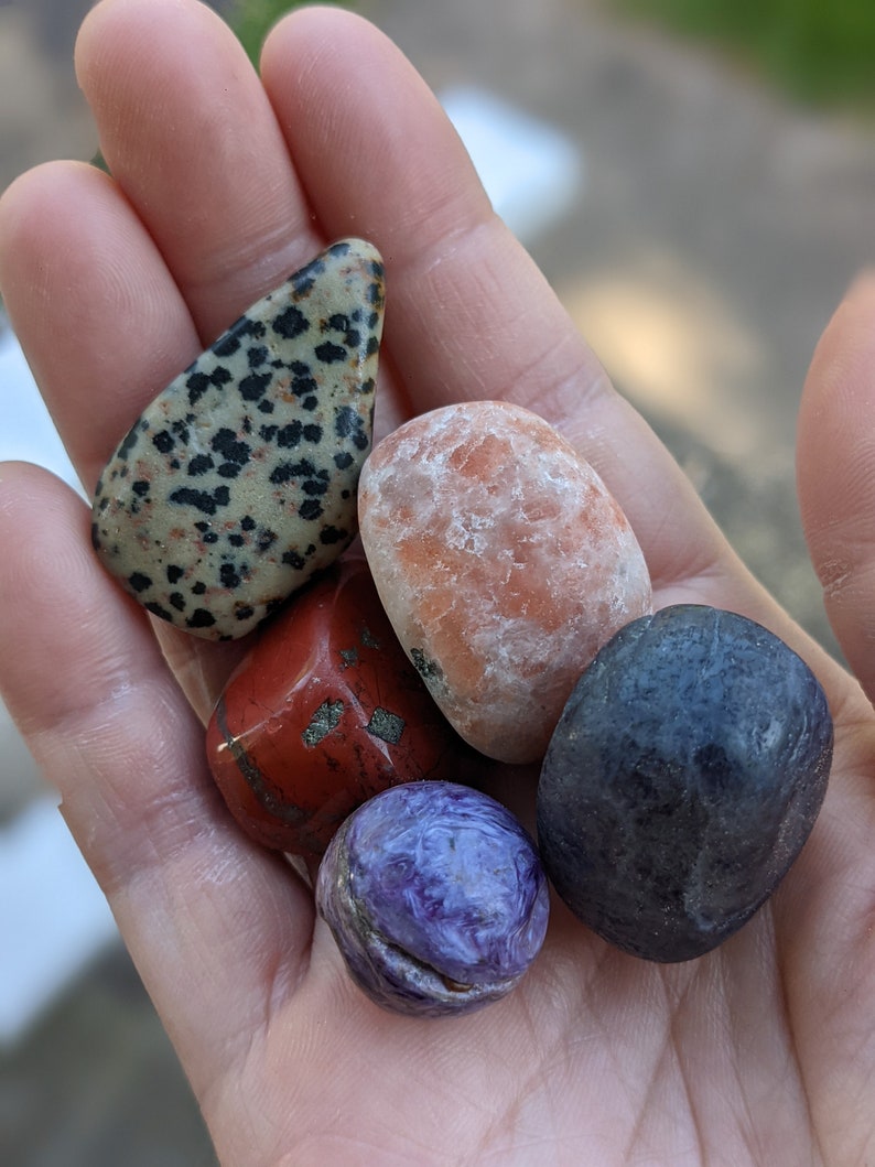 May include: A collection of five polished stones in various colors and patterns, including a black and white speckled stone, a red stone, a light orange stone, a dark blue stone, and a purple stone.