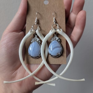 Peut inclure: Une paire de boucles d'oreilles en argent avec un pendentif en agate bleue. Les cercles sont en os et ont une forme délicate et incurvée.