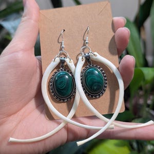 Peut inclure: Une paire de boucles d'oreilles uniques avec des pierres ovales de malachite vert foncé serties dans des montures argentées ornées. Les boucles d'oreilles sont suspendues à des éléments blancs en forme d'os, créant un design distinctif. Présentées sur une carte brune.