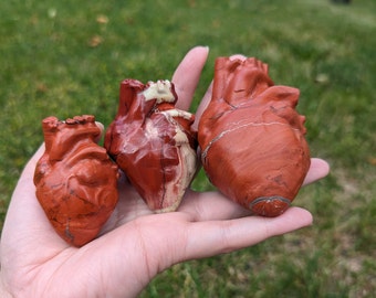 May include: Three red jasper heart-shaped stones, carved and polished, with natural white veining. The stones are held in a hand, with the largest stone in the center.