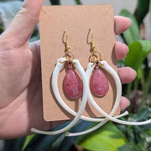 Peut inclure: Une paire de boucles d'oreilles uniques avec des pierres précieuses roses en forme de goutte suspendues à des crochets dorés. Les pierres précieuses sont serties dans un cadre cuivré et entourées de structures blanches et incurvées en forme d'os.