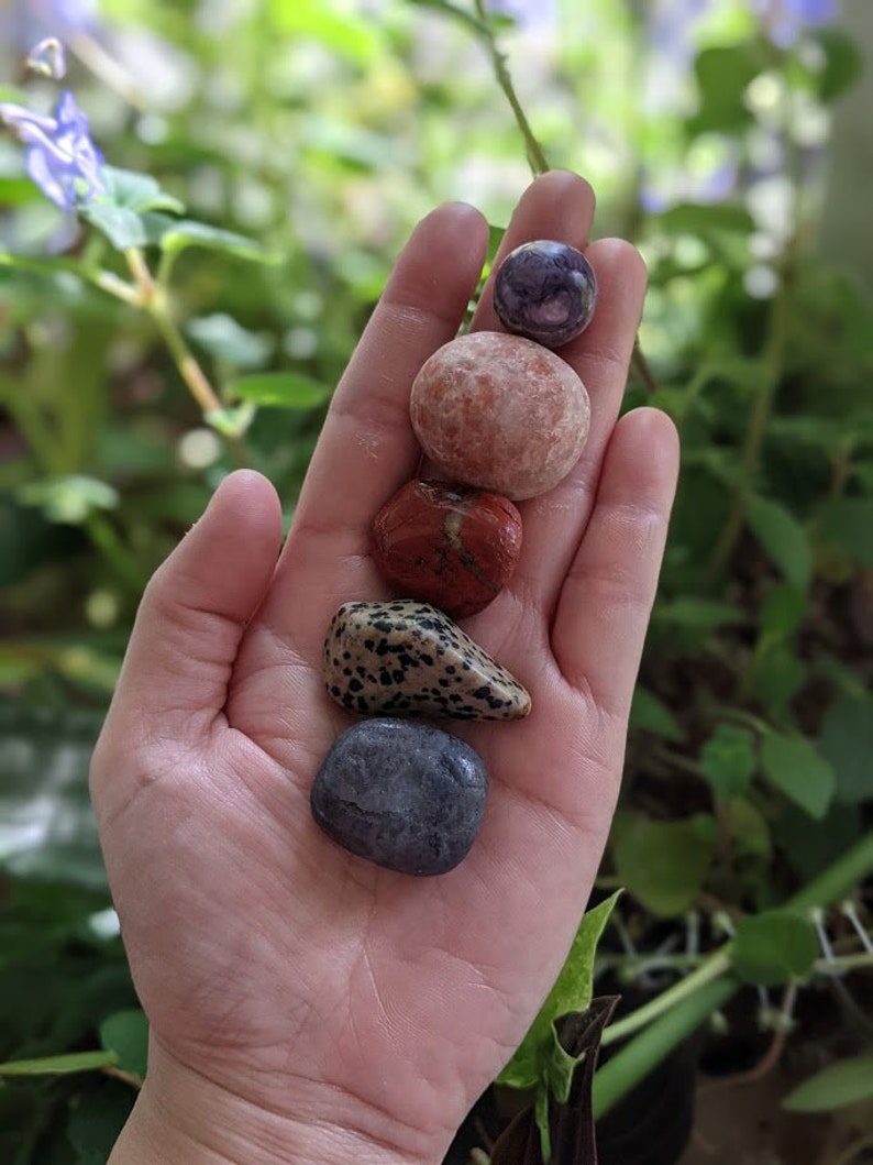 May include: A collection of five smooth, round stones in various colors and patterns, including purple, pink, red, brown with white spots, and gray. The stones are arranged vertically in a hand, with the largest stone at the top and the smallest at the bottom.