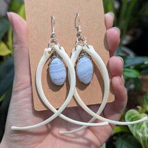 Peut inclure: Une paire de boucles d'oreilles uniques avec des pierres ovales bleu clair serties dans du métal argenté, suspendues à des éléments en forme d'os. Les boucles d'oreilles sont présentées sur un fond neutre, mettant en valeur leur design distinctif.