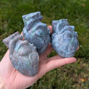 May include: Three anatomical heart sculptures carved from stone with a mottled blue-grey and deep red speckled texture. Each piece features a realistic heart shape with visible chambers and vessels, showcasing a smooth, polished finish throughout.