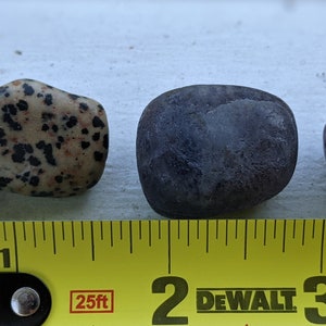 May include: Three smooth stones of different sizes and colors. A speckled brown and black stone, a dark purple stone, and a round purple and white stone. A yellow measuring tape with black markings is below the stones. The tape reads "25ft" and "DEWALT".