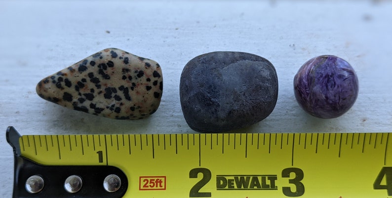 May include: Three smooth stones of different sizes and colors. A speckled brown and black stone, a dark purple stone, and a round purple and white stone. A yellow measuring tape with black markings is below the stones. The tape reads "25ft" and "DEWALT".