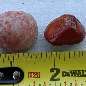 May include: Two red stones, one round and one irregularly shaped, are placed next to a yellow measuring tape. The tape reads "25ft" and has markings for inches and fractions of inches. The round stone is about 1 inch in diameter, and the other stone is about 1 inch long.