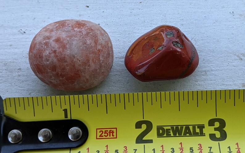 May include: Two red stones, one round and one irregularly shaped, are placed next to a yellow measuring tape. The tape reads "25ft" and has markings for inches and fractions of inches. The round stone is about 1 inch in diameter, and the other stone is about 1 inch long.