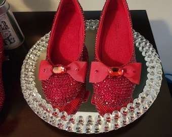 May include: A pair of red sparkly shoes with a red bow and a red jewel in the center of the bow. The shoes are on a silver tray with a clear crystal border.