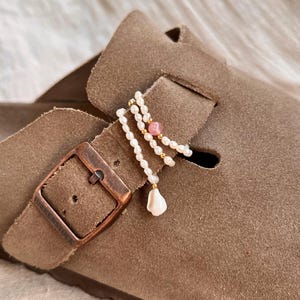 Könnte beinhalten: Ein brauner Wildleder-Clog mit Metallschnalle und einem dekorativen Wickel-Accessoire aus kleinen weißen Süßwasserperlen, winzigen goldfarbenen Perlen, einer einzelnen runden rosa Steinperle und einem kleinen weißen Muschelanhänger.