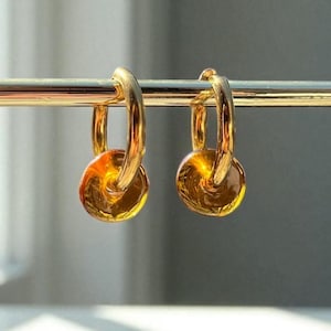 May include: A pair of gold hoop earrings, each featuring a small, amber-coloured disc charm. The earrings are displayed on a gold stand, with a blurred background. The earrings are a stylish accessory.