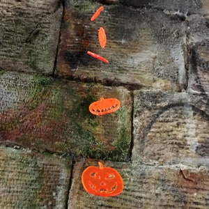 Könnte beinhalten: Zwei orangefarbene Kürbis-Ausschnitte vor einem Hintergrund aus Steinmauer. Die Ausschnitte hängen oben im Bild und überlappen sich leicht.