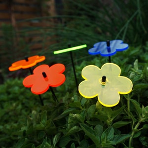 Può includere: Quattro paletti da giardino in plastica a forma di fiore di colore blu, giallo, arancione e rosso. I paletti sono inseriti nel terreno e sono circondati da fogliame verde.
