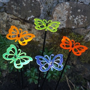 Puede incluir: Cinco estacas de jardín de mariposa de colores vivos hechas de acrílico. Las mariposas son en tonos de verde, naranja, azul y amarillo. Las estacas son de metal negro.