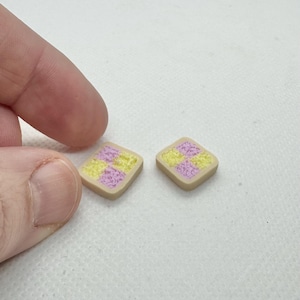Può includere: Due piccoli orecchini quadrati con bordo beige e motivo a scacchi giallo e lavanda. Gli orecchini sono tenuti dalle dita di una persona su sfondo bianco.