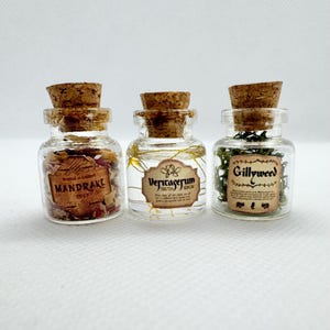 May include: Three small glass bottles with cork stoppers. Each bottle contains different contents and a label. The labels read "Mandrake", "Veritaserum", and "Gillyweed". The bottles are arranged in a row against a white background.