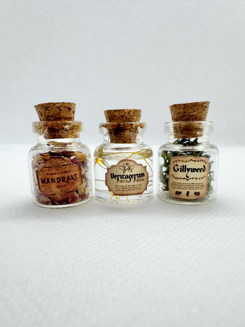 May include: Three small glass bottles with cork stoppers. Each bottle contains different contents and a label. The labels read "Mandrake", "Veritaserum", and "Gillyweed". The bottles are arranged in a row against a white background.