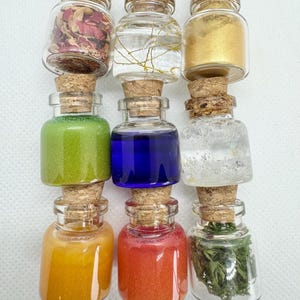 May include: Nine small glass bottles with cork stoppers, each filled with different colored contents. The bottles contain dried flowers, gold glitter, green and blue liquids, and other colorful materials. The bottles are arranged in three rows of three.