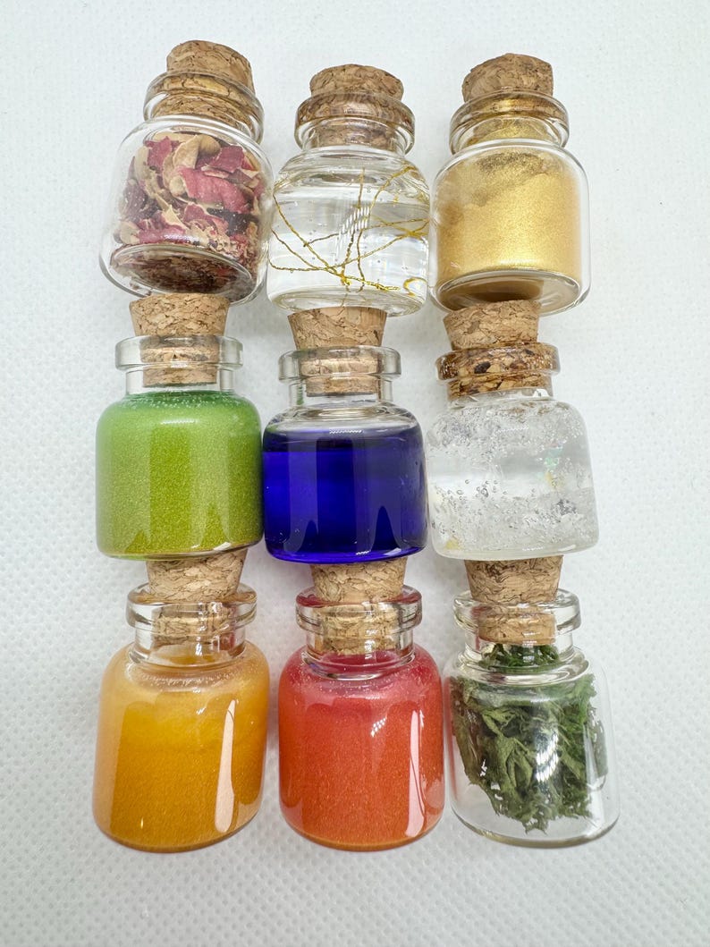 May include: Nine small glass bottles with cork stoppers, each filled with different colored contents. The bottles contain dried flowers, gold glitter, green and blue liquids, and other colorful materials. The bottles are arranged in three rows of three.