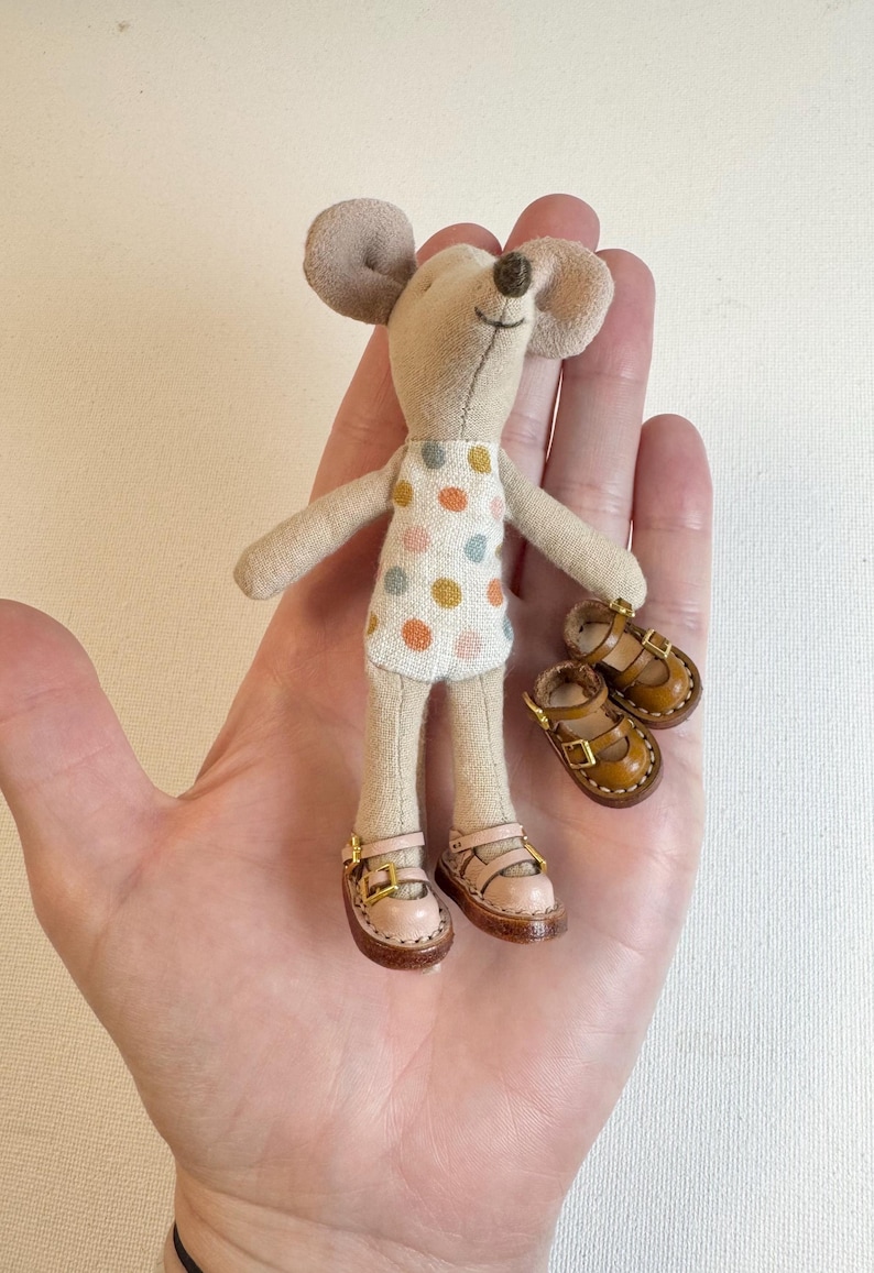 May include: A handmade doll in the shape of a mouse, wearing a polka-dot dress and pink shoes. The doll has tan fabric for its body and large ears. A pair of brown leather shoes with buckles are next to the doll.
