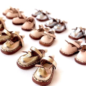 May include: A collection of miniature leather shoes in various colors, including gold, silver, and beige. The shoes have a simple design with a strap across the top and a rounded toe.