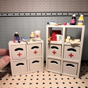 May include: Miniature white medicine cabinets with red crosses and curved drawer pulls. The cabinets are filled with tiny medicine bottles, bandages, and other medical supplies. The scene is set in a dollhouse bathroom.