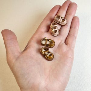 May include: Four miniature leather baby shoes in shades of tan and brown, with buckle closures. Two pairs of shoes are visible, each with a strap across the top and a small heel. The shoes are displayed in the palm of a hand.
