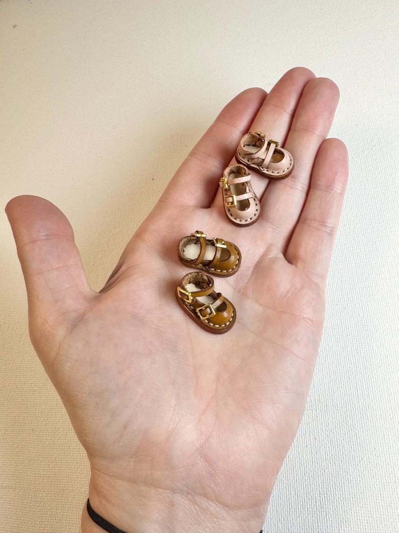 May include: Four miniature leather baby shoes in shades of tan and brown, with buckle closures. Two pairs of shoes are visible, each with a strap across the top and a small heel. The shoes are displayed in the palm of a hand.