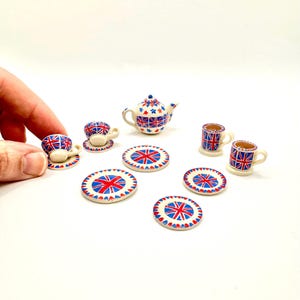 May include: A miniature tea set featuring a teapot, cups, saucers, and plates. The set is decorated with a red, white, and blue Union Jack flag design. The teapot has a blue handle and spout. The plates and saucers are round.