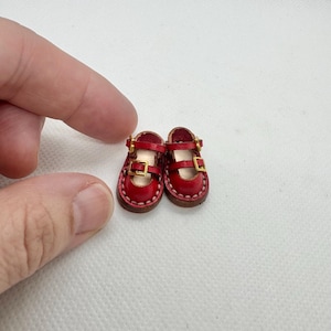 May include: A pair of miniature red Mary Jane shoes with gold-colored buckles. The tiny shoes have a stitched brown sole and are designed for a doll or other small figure. The shoes are made of leather.