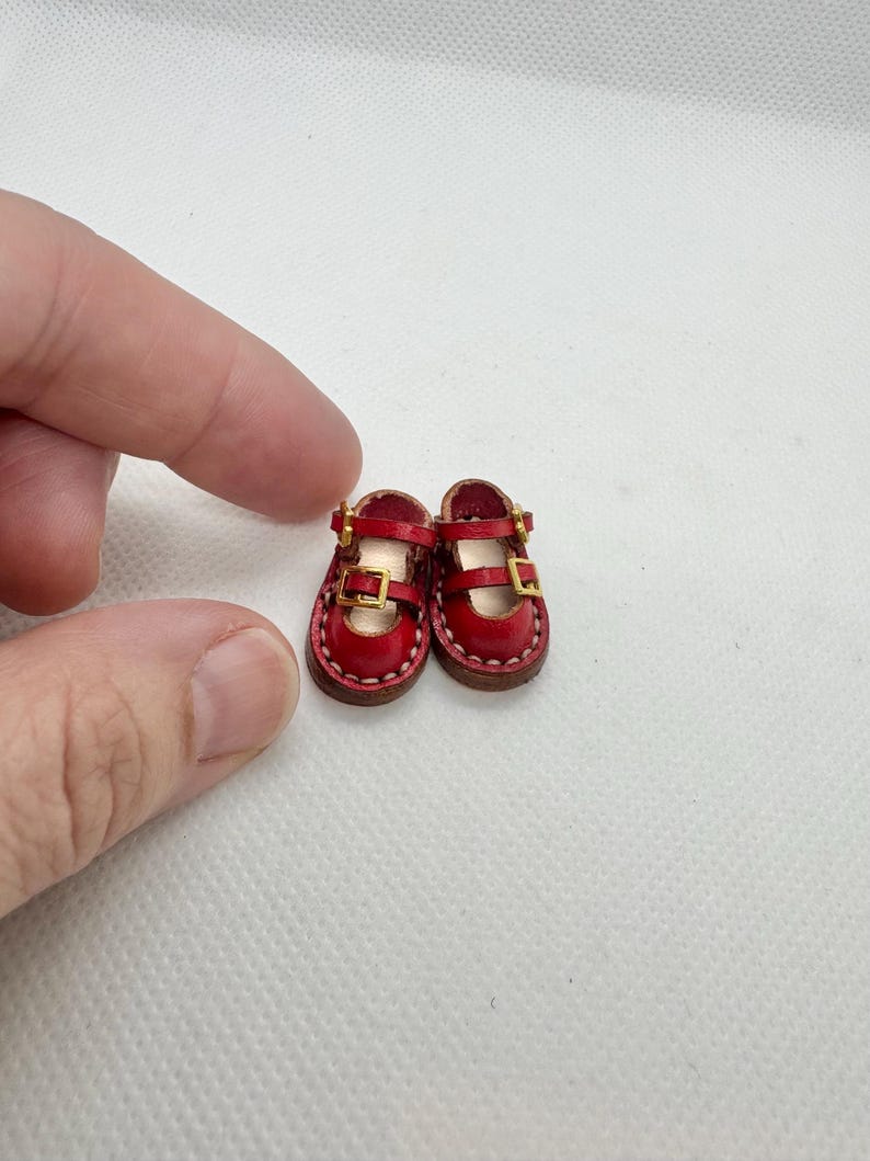 May include: A pair of miniature red Mary Jane shoes with gold-colored buckles. The tiny shoes have a stitched brown sole and are designed for a doll or other small figure. The shoes are made of leather.