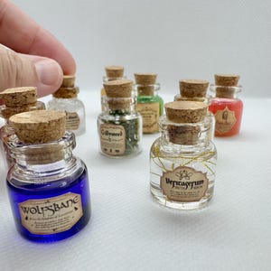 May include: A collection of miniature glass potion bottles with cork stoppers. The bottles contain various colored liquids and herbs, with labels such as "Wolfsbane" and "Veritaserum". The bottles are arranged on a white surface.