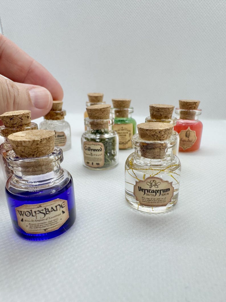 May include: A collection of miniature glass potion bottles with cork stoppers. The bottles contain various colored liquids and herbs, with labels such as "Wolfsbane" and "Veritaserum". The bottles are arranged on a white surface.