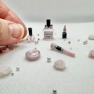 May include: A miniature set of pink and black beauty products, including a perfume bottle with the word "PARFUM" on it, a lipstick, a small bottle of perfume, a compact with a pink bow, and a makeup brush.
