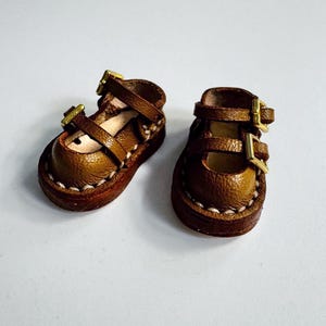 May include: A pair of miniature brown leather Mary Jane shoes with gold-colored buckles. The shoes have a rounded toe and are stitched with white thread around the sole. The buckles are rectangular and secure the straps.