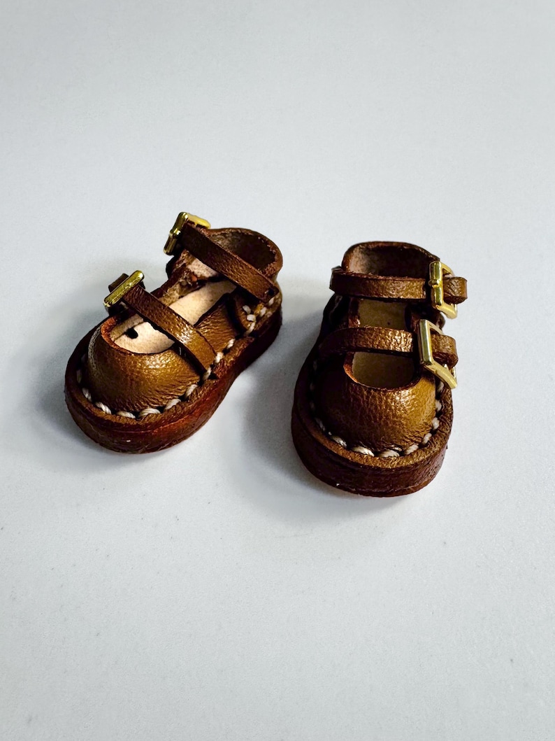 May include: A pair of miniature brown leather Mary Jane shoes with gold-colored buckles. The shoes have a rounded toe and are stitched with white thread around the sole. The buckles are rectangular and secure the straps.