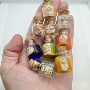 May include: A collection of miniature potion bottles with cork stoppers. The bottles contain various colored liquids and powders, with labels such as "Floo Powder" and "Felix Felicis". The bottles are held in a hand.