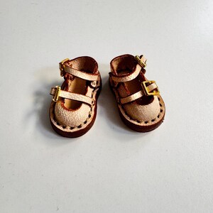 May include: A pair of miniature leather Mary Jane shoes. The shoes are tan with brown soles and straps, featuring gold-colored buckles. The edges are stitched with dark brown thread, and the shoes appear to be handmade.