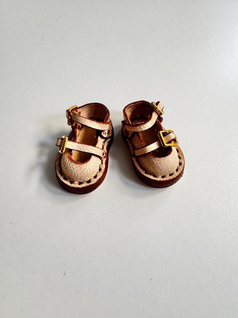 May include: A pair of miniature leather Mary Jane shoes. The shoes are tan with brown soles and straps, featuring gold-colored buckles. The edges are stitched with dark brown thread, and the shoes appear to be handmade.