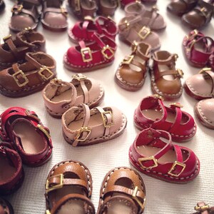 May include: A collection of miniature leather shoes in various colors, including brown, pink, and red. The shoes feature buckle closures and are designed for dolls or crafts. The image showcases the variety of styles and colors available.