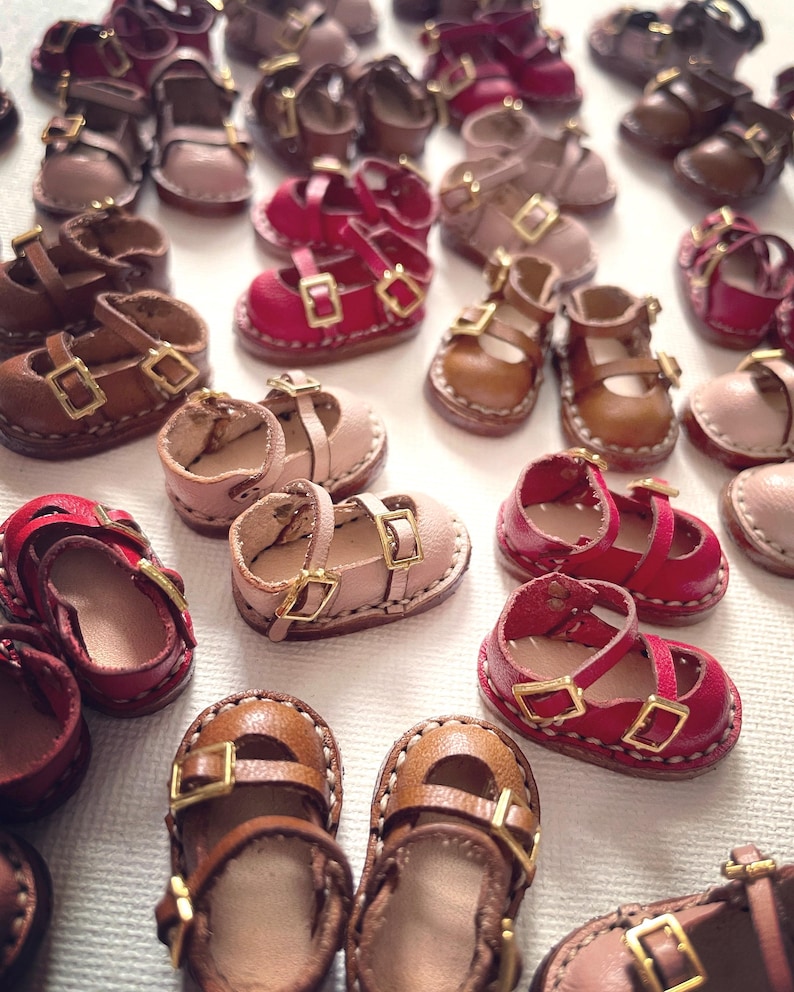May include: A collection of miniature leather shoes in various colors, including brown, pink, and red. The shoes feature buckle closures and are designed for dolls or crafts. The image showcases the variety of styles and colors available.